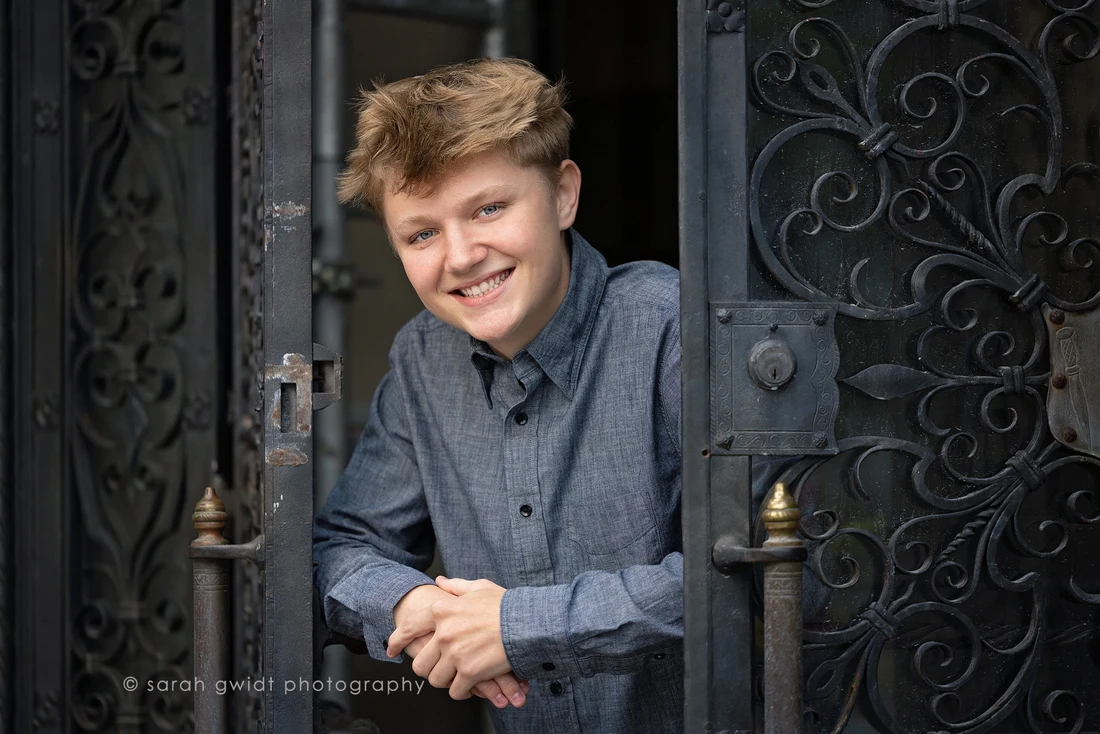 Jace smiling in a blue-gray shirt beside an ornate doorway; photograph by Sarah Gwidt Photography